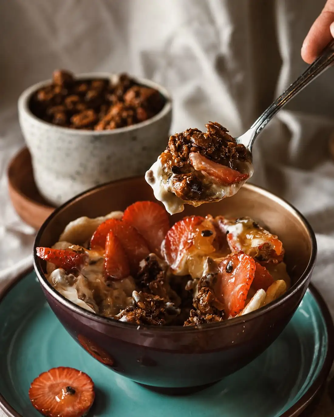 A bowl of yoghurt, granola and sliced strawberries with a spoon lifting a mouthful.