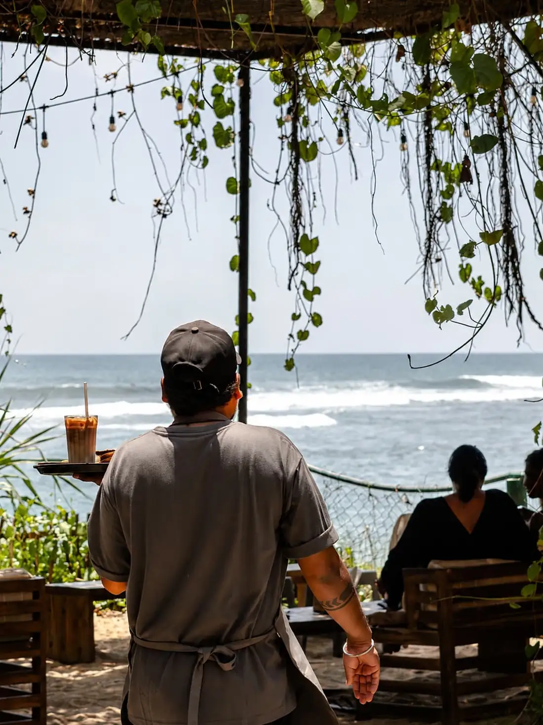 A member of the Kāffi team carrying an iced coffee on a tray towards the open front of the café, with the sea and breaking waves beyond.