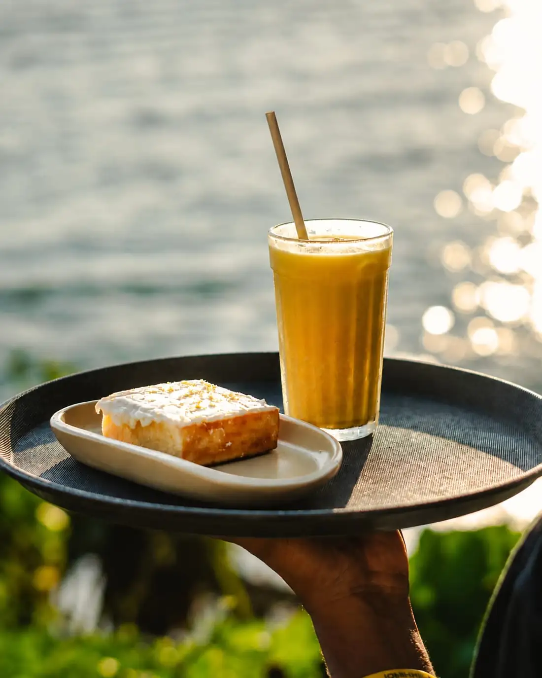 A tray carried through the garden at golden hour, holding a lemon slice and a mango smoothie against the water.