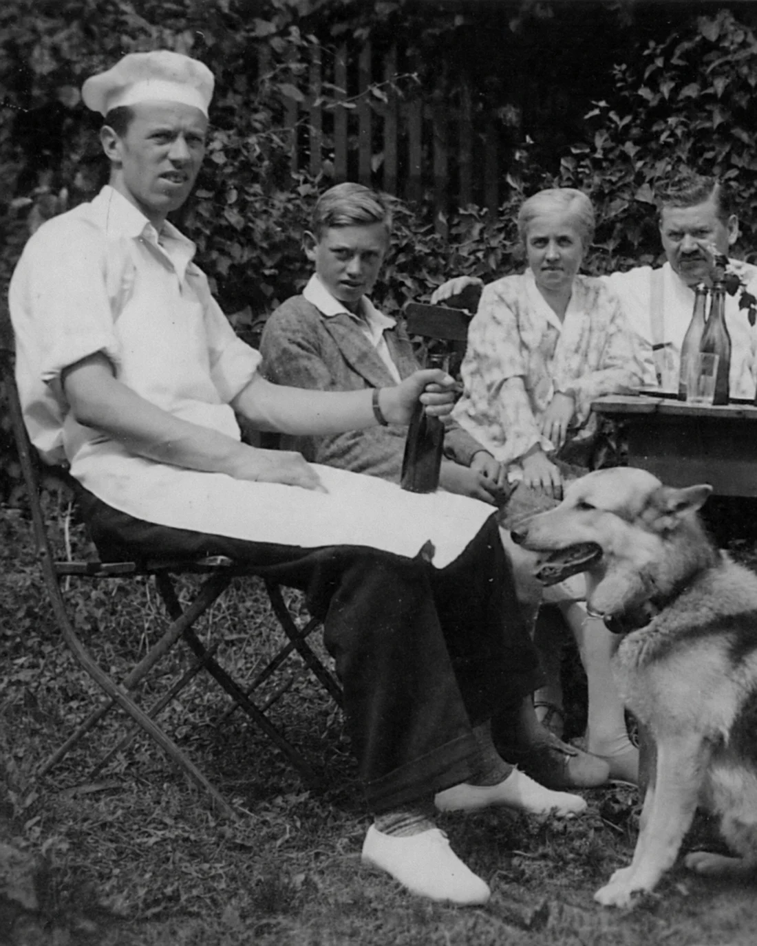 Black-and-white family photograph from the 1930s showing a baker in white seated in a garden with relatives and two dogs.
